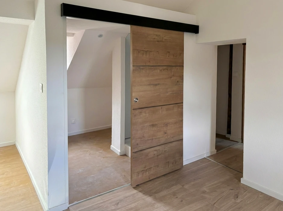 Intérieur d'un appartement sous combles vide, porte coulissante type barn door en bois massif aspect chêne vieilli, montée sur rail métallique noir en applique
