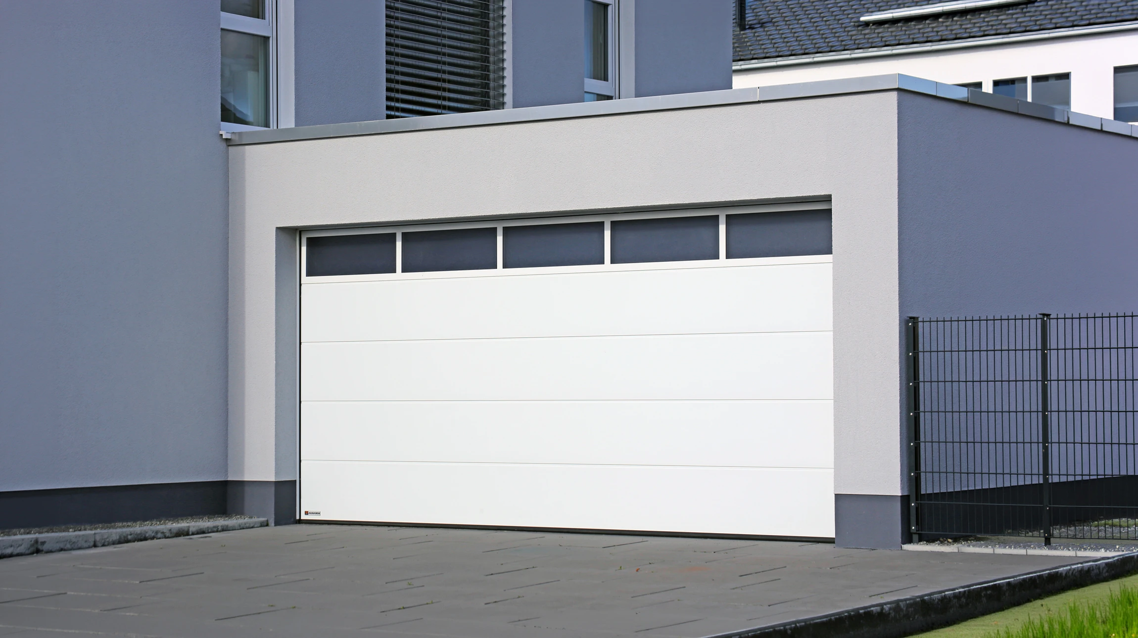 Technicien en tenue professionnelle (salopette ou combinaison) effectuant une réparation ou entretien sur une porte de garage enroulable ou basculante, en train de travailler sur le mécanisme, les ressorts ou la motorisation. La porte de garage est clairement visible en arrière-plan. La scène se déroule devant le domicile ou garage d'un client particulier, en plein jour. L'image doit transmettre la confiance, la compétence et la proximité avec le client particulier.