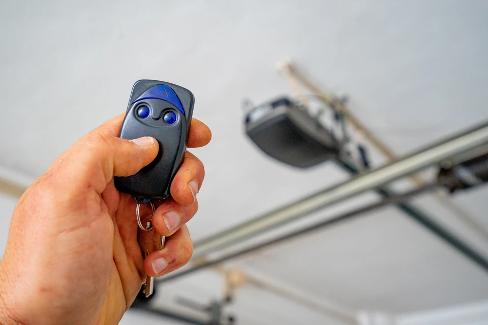 Un technicien qualifié en tenue de travail, accroupi ou debout près d'une porte de garage, effectuant une réparation ciblée (détail visible du ressort, du moteur, du câble ou du rail en cours de réparation). L'arrière-plan montre une habitation résidentielle. La scène illustre une intervention de réparation efficace et professionnelle, sans remplacement de la porte.